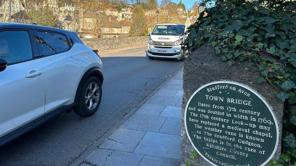 The bridge, with a couple of vehicles crossing, and the plaque detailing it's history