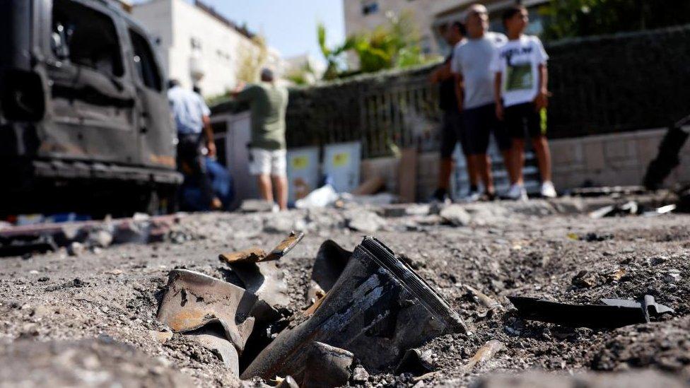 Metal remains of rocket embedded in the ground. People in the background look on.