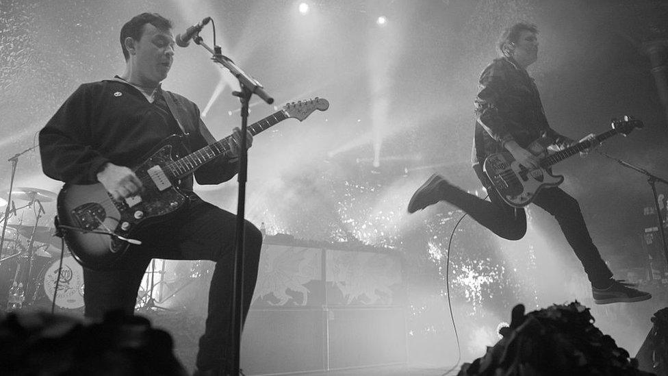 Manic Street Preachers performing, with bassist Nicky Wire leaping in the air and lead singer James Dean Bradfield at the mic with guitar