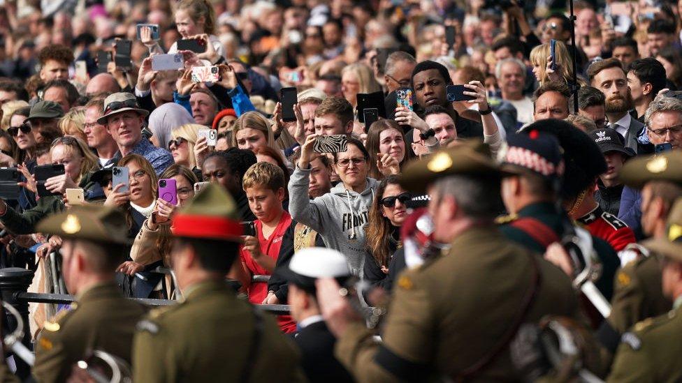 Large crowd of people, holding their phones as the military goes past.