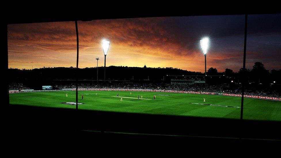 The light towers of Manuka Oval are lit up in front of the sunset.