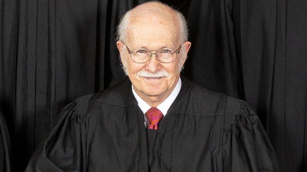 Tom parker wearing a black robe and glasses, sitting in front of a black backdrop