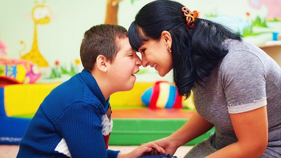 Young boy in blue top smiling and holding his head against the head of a woman, who is also smiling