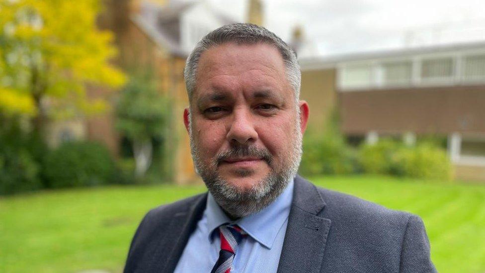 Man with short dark hair and beard wearing a suit and tie and standing outside