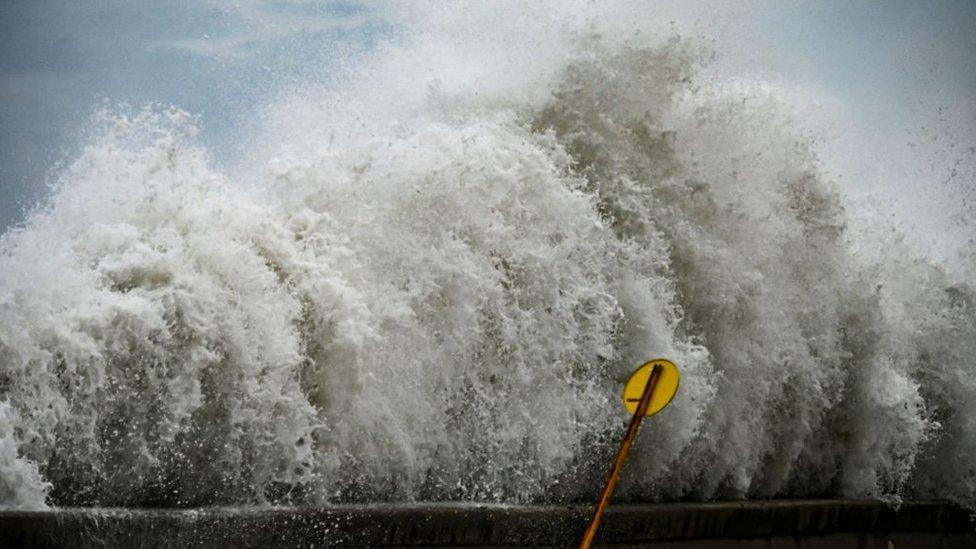 Large amounts of sea foam crashes up against a barrier knocking over a sign.