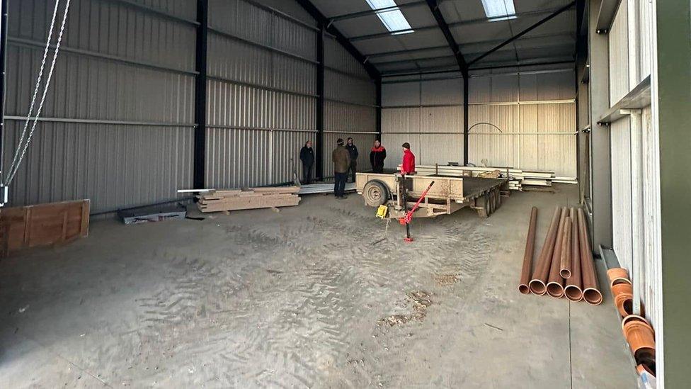 Large mostly-empty warehouse-like building with copper pipes and planks of wood. Five people are waiting to start work.