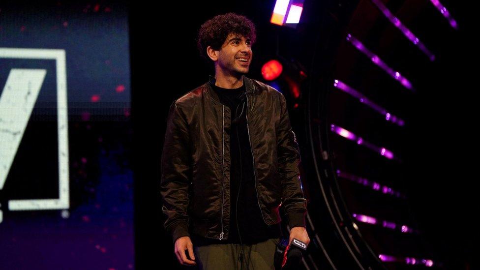 Tony Khan, co-founder of AEW, pictured on stage. Tony, a man in his 30s, has short curly dark hair and brown eyes. He is wearing green trousers and a black bomber jacket over a black T-shirt. In his left hand he is holding a microphone and is smiling off stage to the right of the camera.