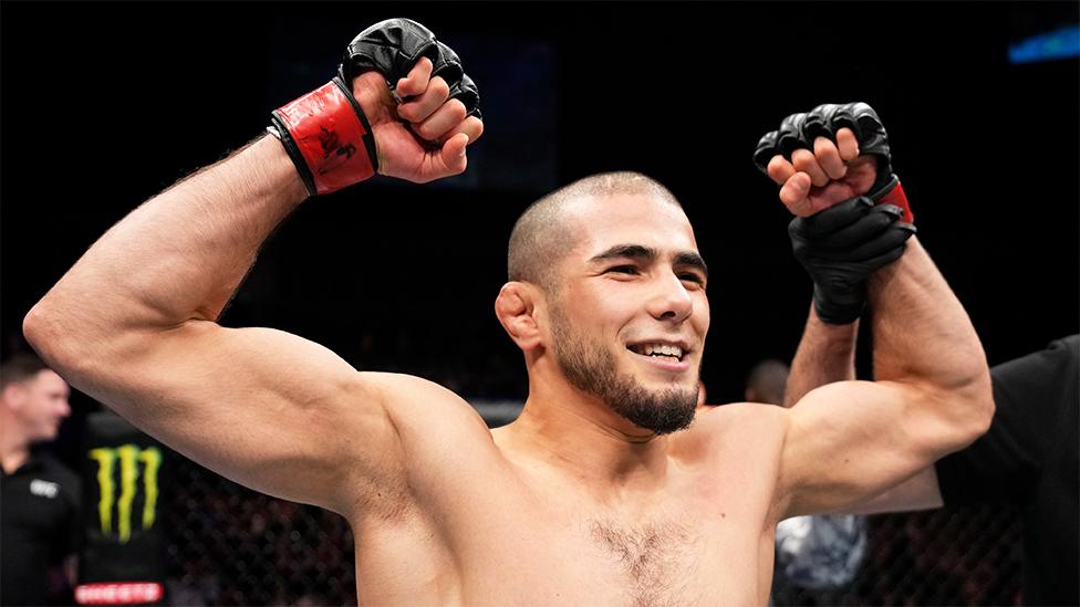 Muhammad Mokaev, a man, celebrates his victory over Cody Durden in a flyweight fight. He is smiling and has both arms in the air. He is wearing black fingerless fighting gloves.