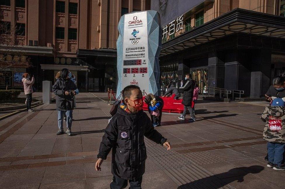 Parents with their children wearing face masks stand next to a countdown display showing days left for the Beijing 2022 Winter Olympics, on a shopping street in Beijing, China, on 17 January 2022