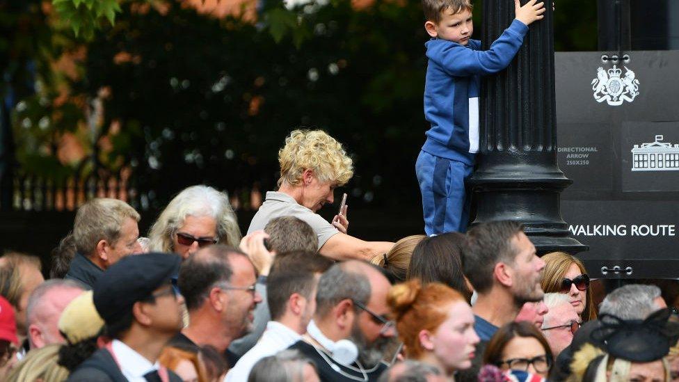 A young boy climbs up onto a lamppost to get a view of the procession, above a crowd of people.