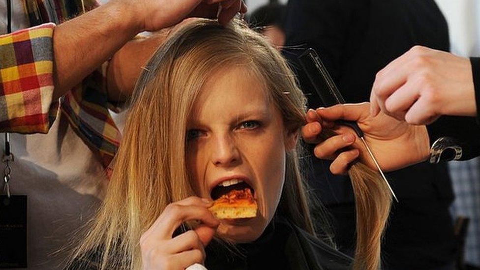 A model takes a bite of pizza while she gets her hair done backstage before the Donna Karan show