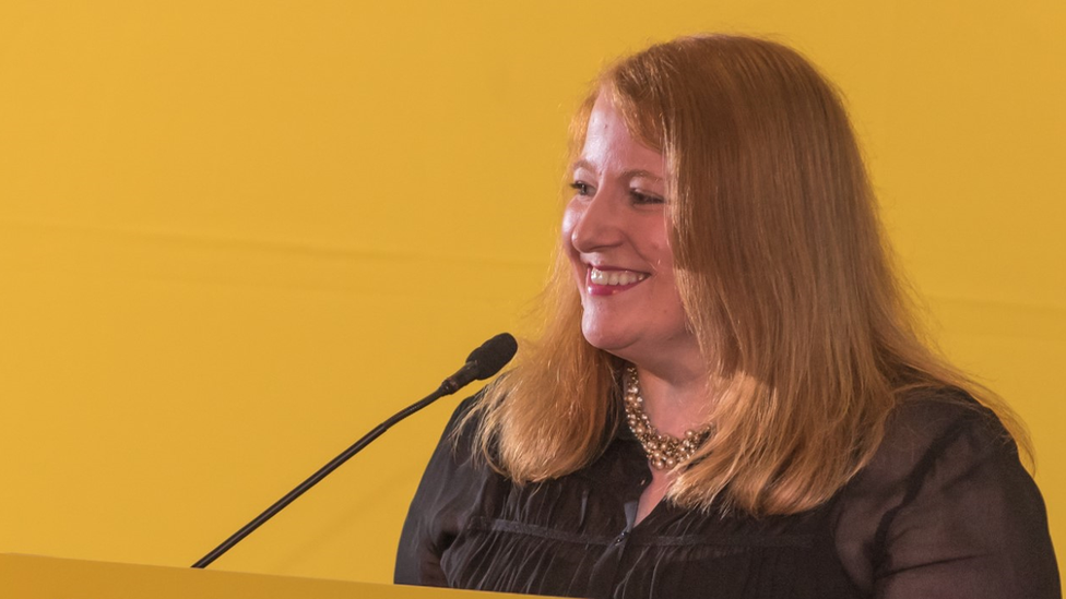 Naomi Long smiles as she speaks into a microphone against a yellow background