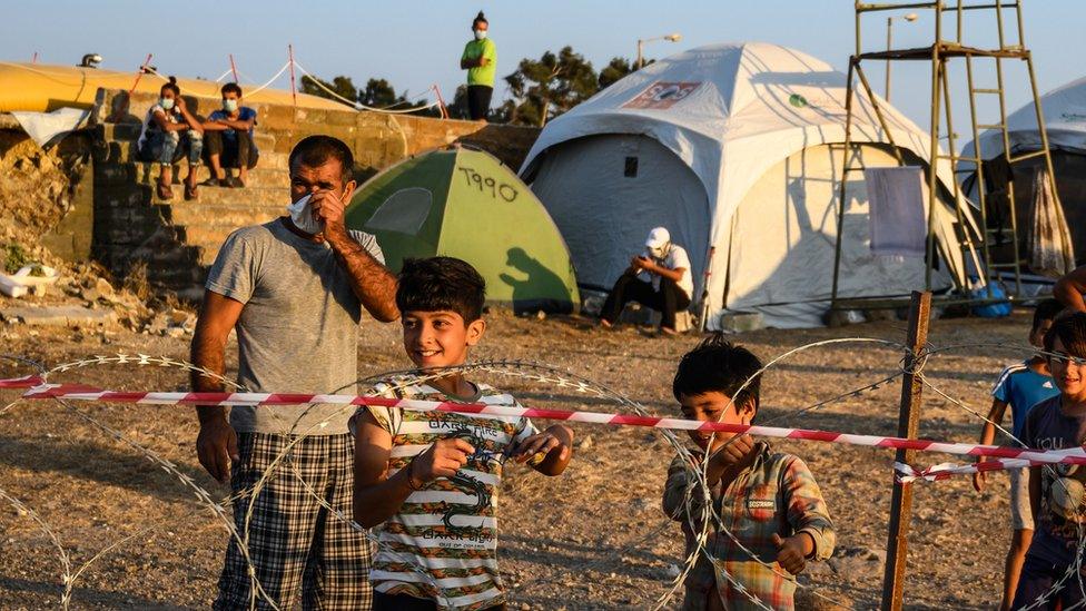 Adults and minors refugees and migrants stand behind razor wire at Kara Tepe camp