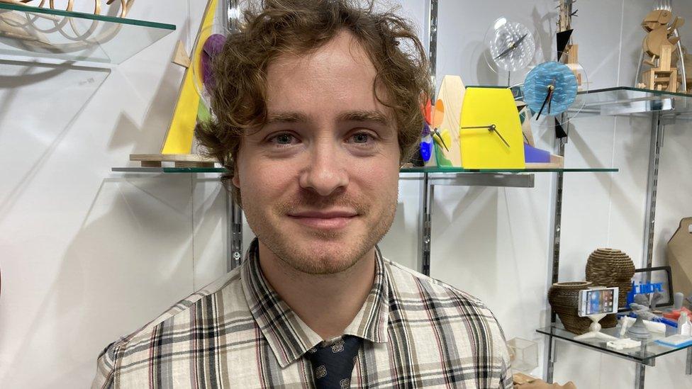 A man in a checked shirt and tie with curly hair in front of some art work