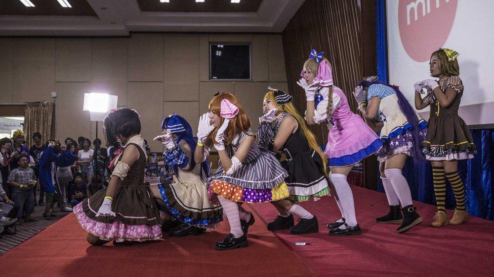 Seven women in cosplay dresses, line up behind one another, in pose, on stage