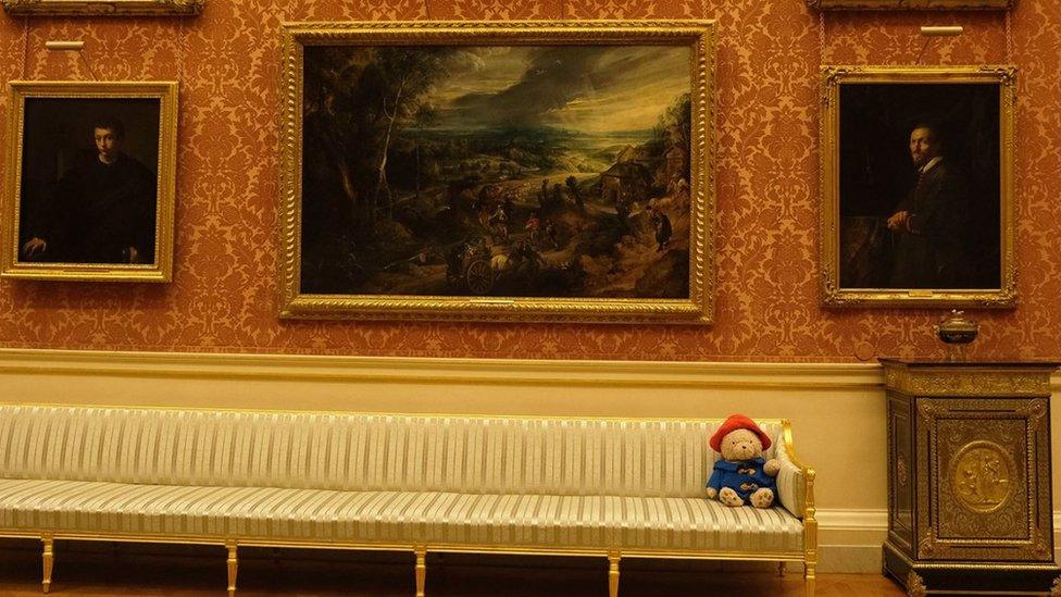 Paddington Bear on a sofa in front of several paintings