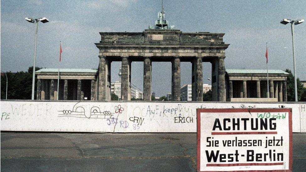 A sign in front of the Berlin Wall and Brandenburg Gate reads "Achtung! Sie verlassen jetzt West-Berlin" (lit. "Attention! You are leaving West Berlin")