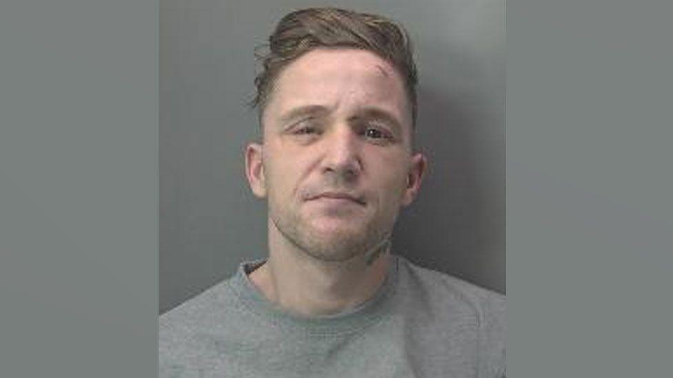 A police mug shot of Dillon Currie aka Abeeb Hussain. He has light brown hair which is longer on the top and very short on the sides and has subble. He is wearing a grey sweatshirt.  