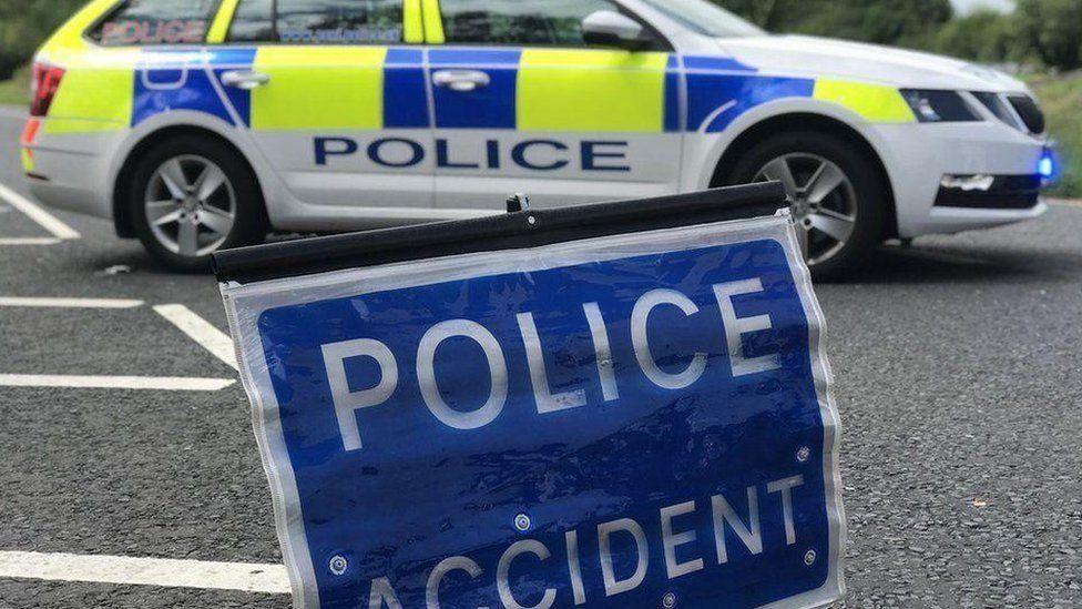 A generic police car parked on a road behind a blue road sign that reads "Police, accident" in bold white writing.