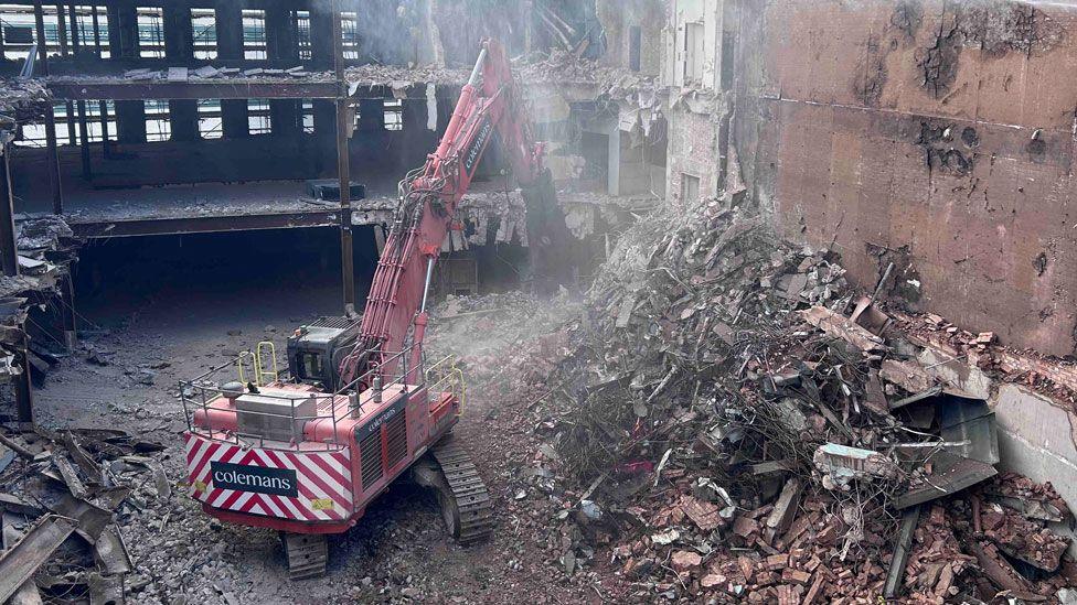 An orange excavator with a large jointed arm at a right angle as seen in the middle of the part-demolished M&S building in Northampton. Its grabber is reaching beyond a pile of rubble on the right to pull at the three storey building, which is open to the elements.