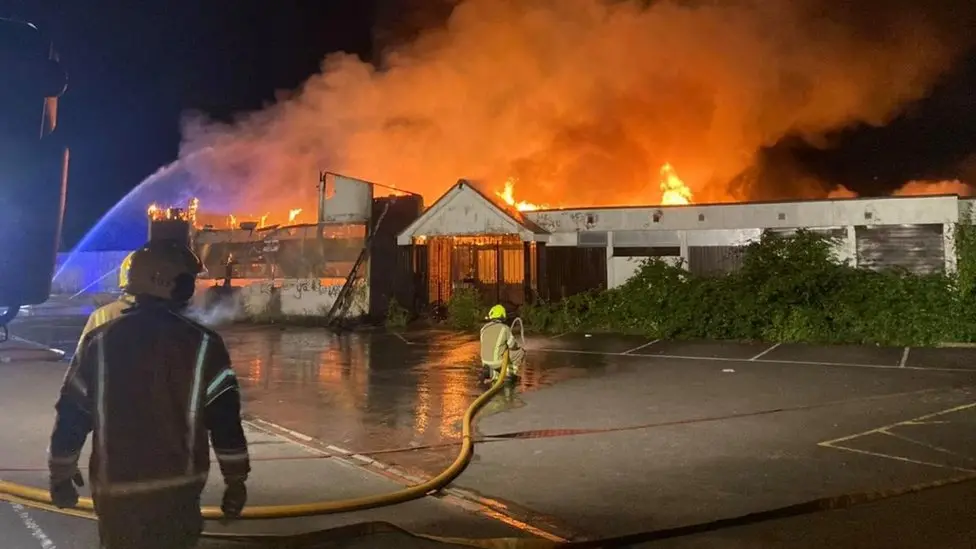 The club's clubhouse is well alight, with fire pouring from the single-storey building, with a firefighter spraying water from a hose close to the building, as another firefighter watches on 