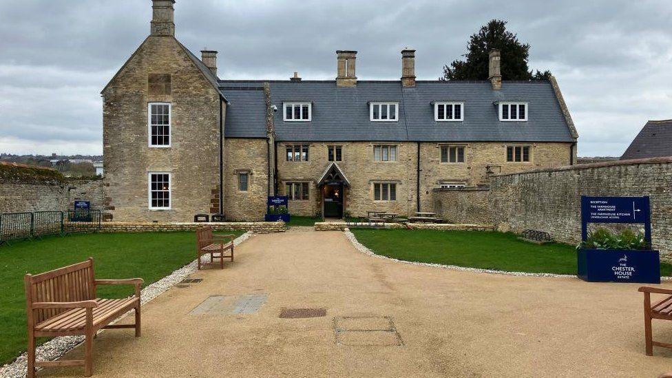 Chester House - a three-storey Northamptonshire stone manor house with gravel path and benches in front. There are three wooden benches, signs and grass areas. 