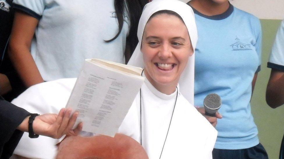 Sr Clare Crockett is sitting, dressed in a habit, while reading from a book being held by one of four young women standing around her.