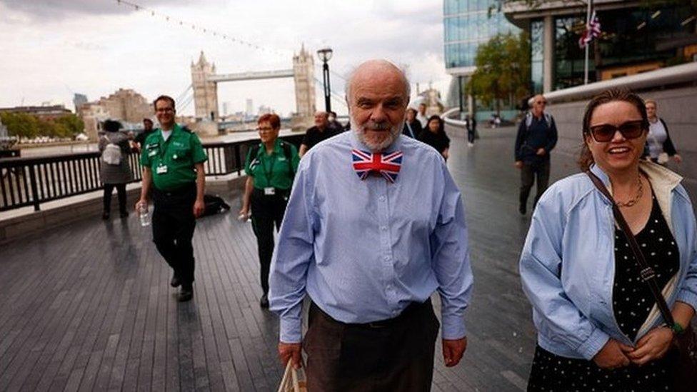 A man in a UK bowtie in the foreground, with two volunteers behind.