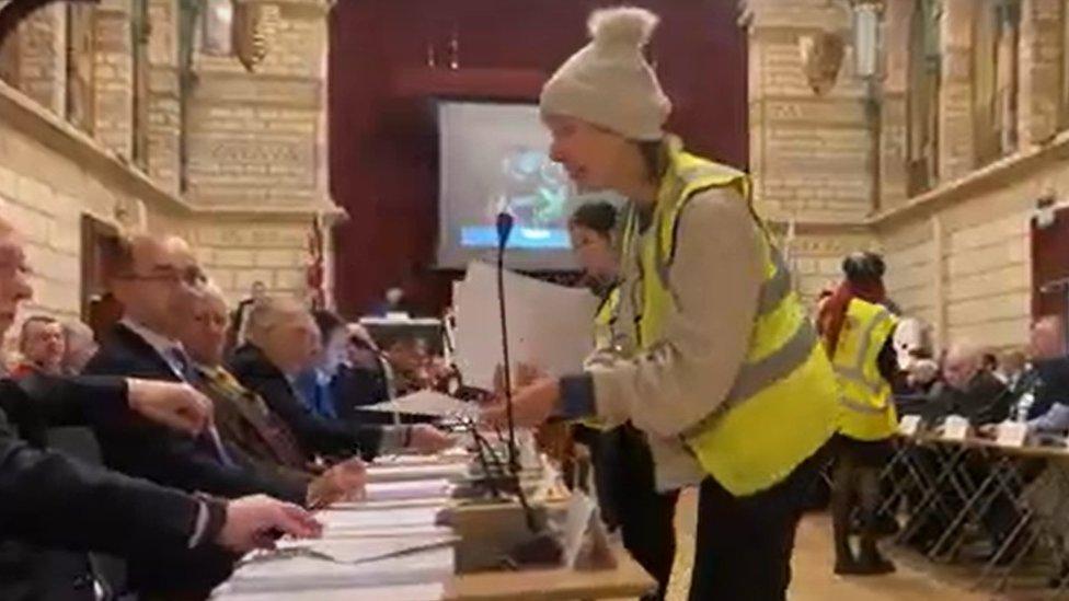 People wearing hi-viz jackets walk amongst tables where councillors are sitting handing out sheets of paper