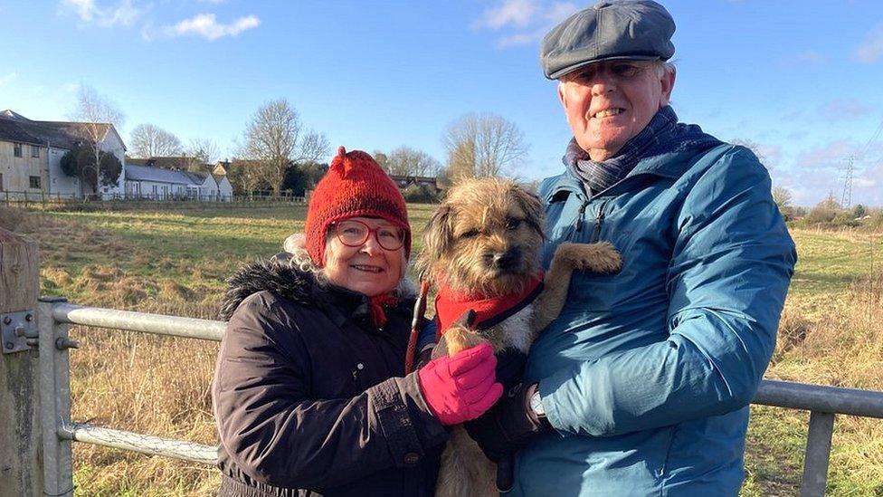 Leila and Mike standing looking at the camera holding their dog between them in winter clothes, Leila in a red woolly hat, Mike in a flat cap in front of a field