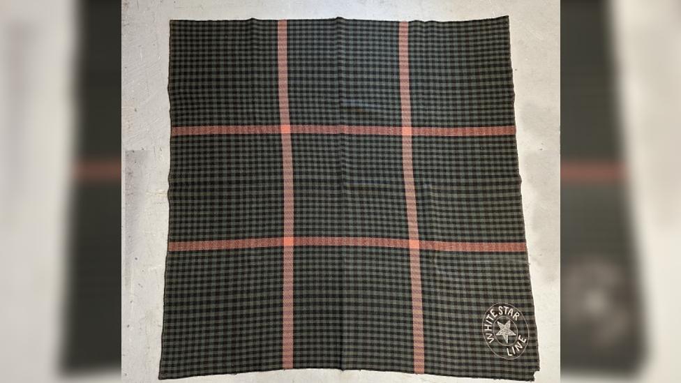 A green/brown/tartan blanket with a red checked pattern with the White Star Line logo in the bottom right corner.