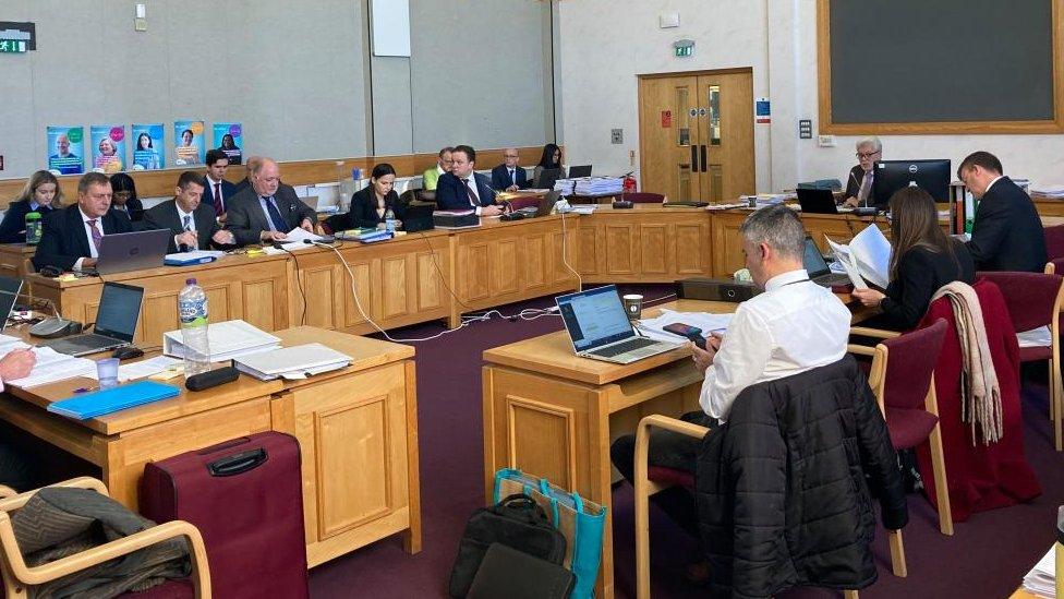 People wearing suits sit around big wooden desks in a council chamber