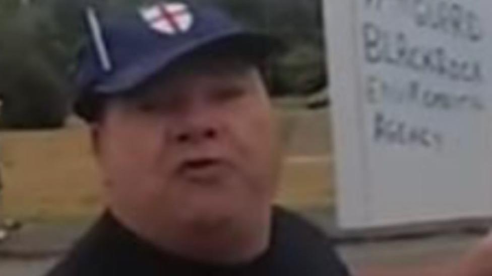 Police image of a man looking angry and wearing a dark blue cap with an England flag logo on it