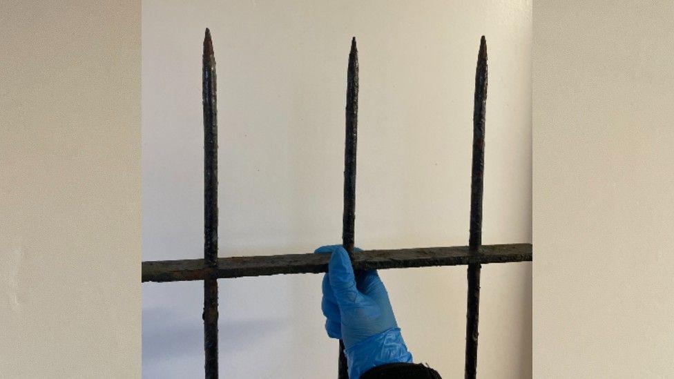 A blue gloved hand holding up a section of black metal fence with a narrow sharp end