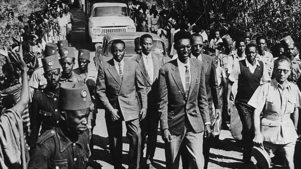 King Kigeli V of Rwanda (front) is flanked by guards and crowds as he is escorted through a town, circa 1959