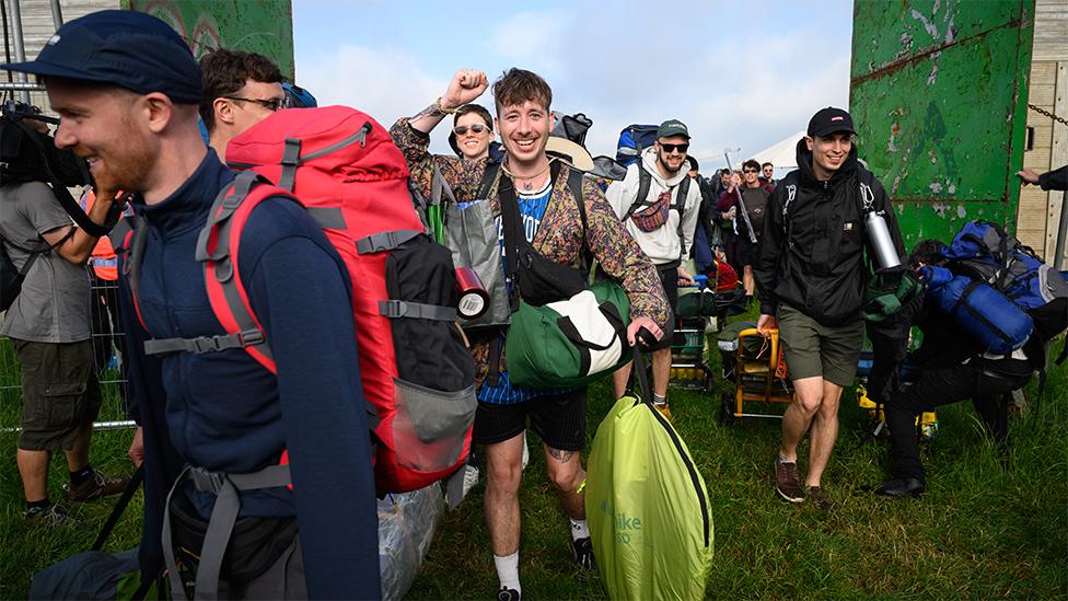 Festival attendees pass through the gates. A man is smiling looking at the camera. Many people have got backpacks on, wearing sunglasses. The gates are coloured green, with a light blue sky.