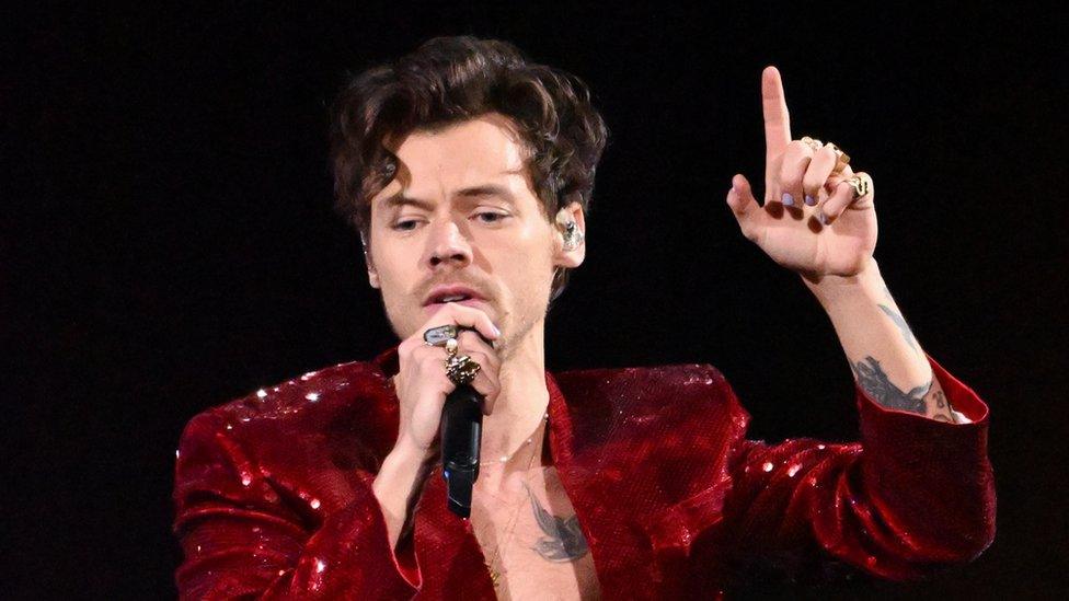 Harry Styles performs at the Brit Awards. He's wearing a glittery burgundy jacket, unbuttoned, which shows off part of his torso. He has one hand help up by his head the index finger pointing upwards. His nails are painted in pastel colours. The other hand is holding a microphone in front of his mouth. He wears gold rings on his fingers.