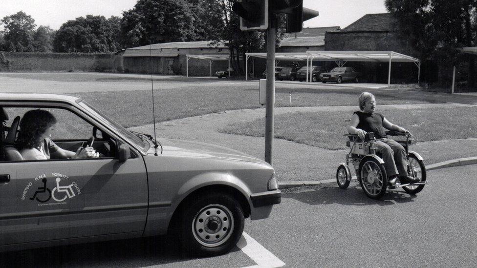 A man using an electric wheelchair crossing the road. A car with mobility logos on the side is parked at the lights