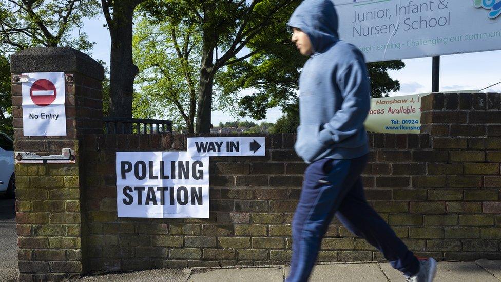 A person, blurred in motion, runs past a sign that uses arrows to point the way towards the nearest local polling station as voters head to polling stations to cast their ballots in local elections