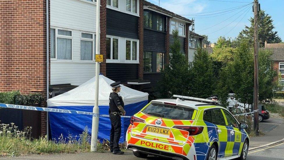 A police car parked outside a house, cordoned off by police tape. There is a blue walled tent, with white roof at the front of the house. A police officer stands next to the car.