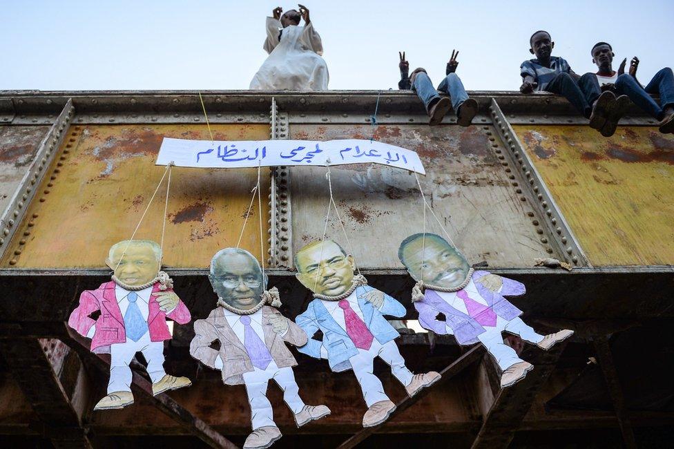 Sudanese men sit together atop a bridge dangling paper puppets showing the faces of the old regime