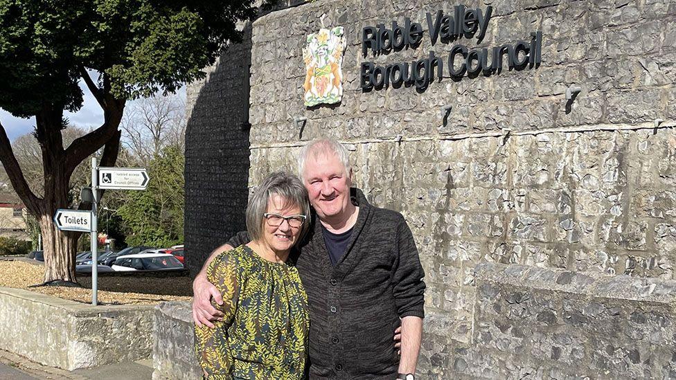 Linda and Alan Boyer smiling below a Ribble Valley Borough Council sign in the sunshine, both are smiling and Alan has his arm round his wife