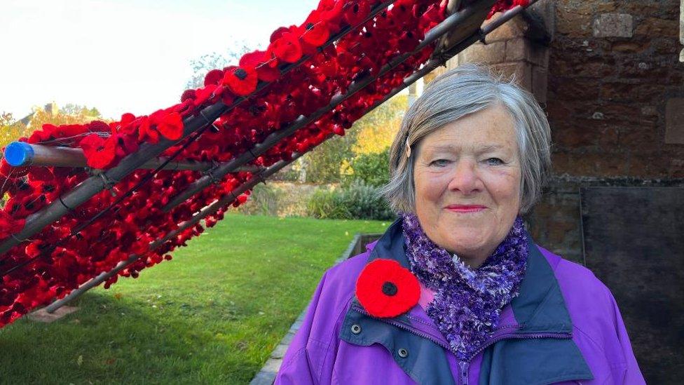 Woman with grey hair wearing a big poppy and a purple coat stands alongside a line of poppies