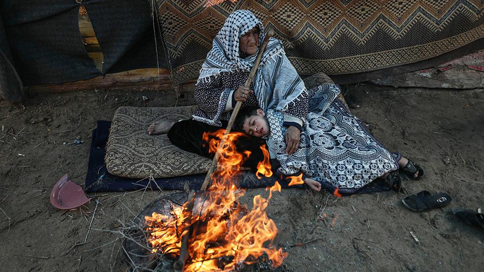 An old woman and a child try to stay warm by burning brushwood in front of a tent house, in Gaza City, in December 2022 - the woman has her head covered in a chequered shawl and is holding a large stick. The boy, with short dark hair, is lying with his head on her lap under a white and grey patterned blanket.