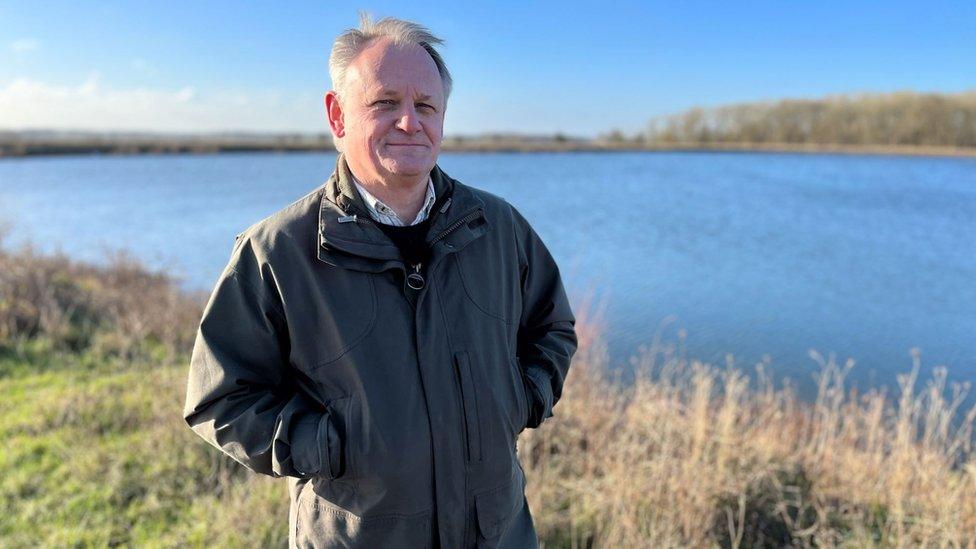 Man with grey hair wearing a green jacket and standing by an expanse of water