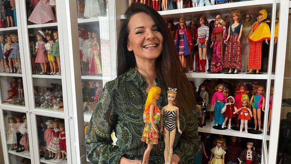 Dawn Austin holding two Barbie dolls, with more of her collection on display behind her