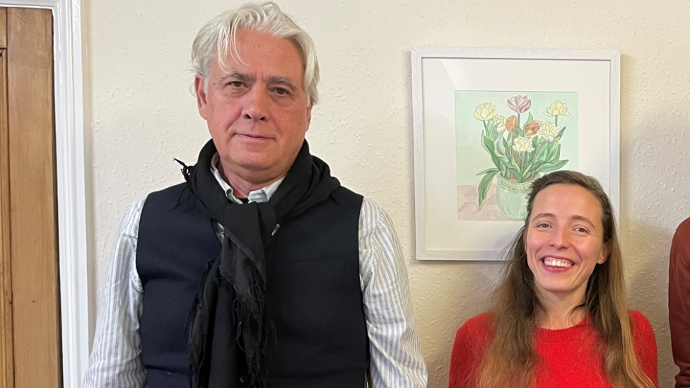 Igor and Kateryna stood against a wall. Igor has short white hair and is wearing a black gilet. Kateryna has long light brunette hair and is wearing a red top. 