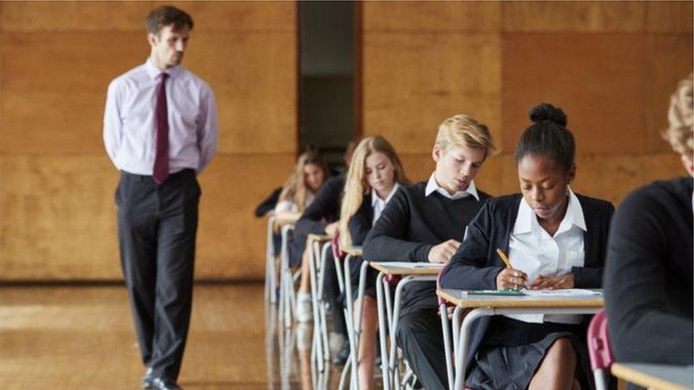 School students sitting an exam in a hall while being monitored by a male adult