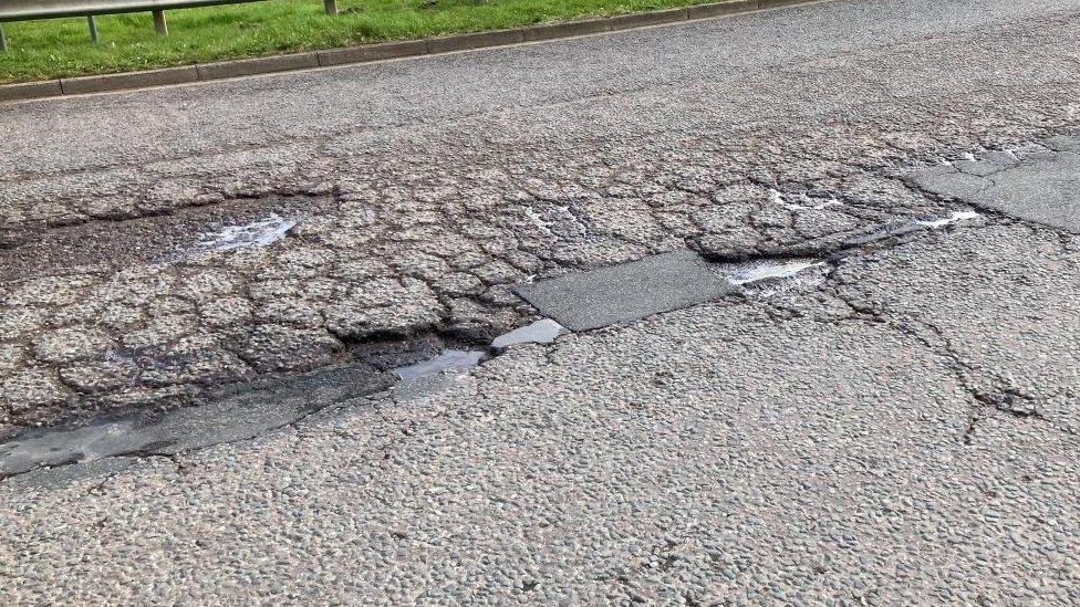 Section of road surface, showing cracks and small water-filled holes