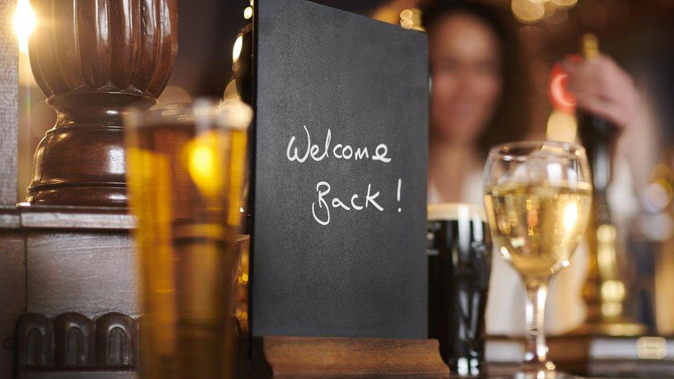 Sign in pub saying welcome back - with out of focus woman behind bar, and drinks on bar